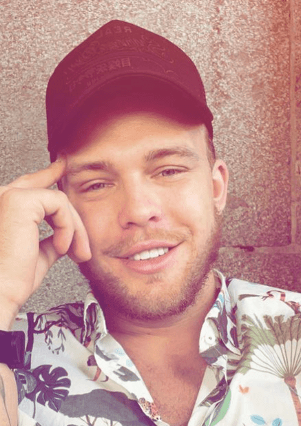 Person wearing a black cap and floral shirt with partially visible necklace against a stone wall background.