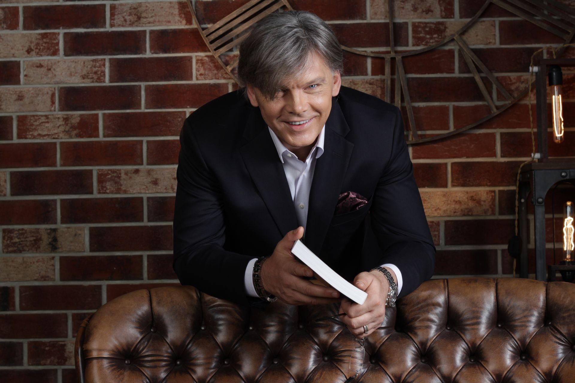 Man in a suit holding a book, leaning over a leather couch with brick wall background.