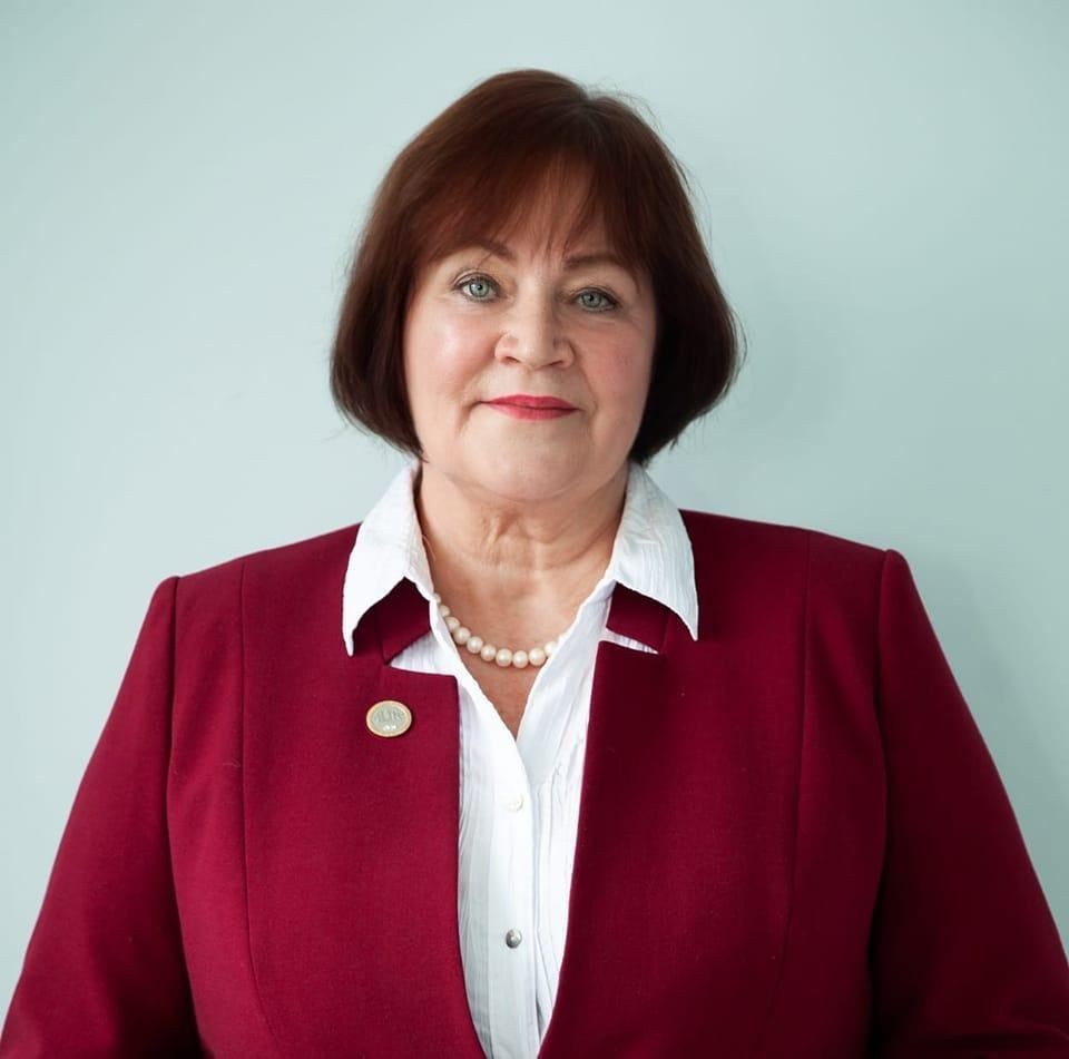Woman in a burgundy blazer with pearl necklace, smiling against a light background.
